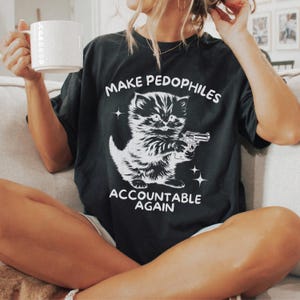 Op de afbeelding: Zwart T-shirt met een witte afbeelding van een katje met een pistool en de tekst "MAKE PEDOPHILES ACCOUNTABLE AGAIN". Het shirt is donker van kleur en heeft een losse pasvorm.