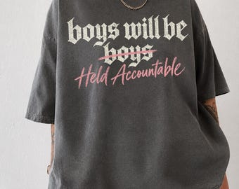 May include: A charcoal gray short-sleeve t-shirt featuring the white text "boys will be boys" with a pink line through the word "boys" and the pink script text "Held Accountable" underneath. The shirt has a crew neckline and a relaxed fit.