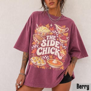 May include: A berry-colored oversized t-shirt featuring a graphic print of various food dishes, including a bowl of mashed potatoes, a casserole, a gravy boat, and a pie. The text reads THE SIDE CHICK in large, bold, cream-colored letters.