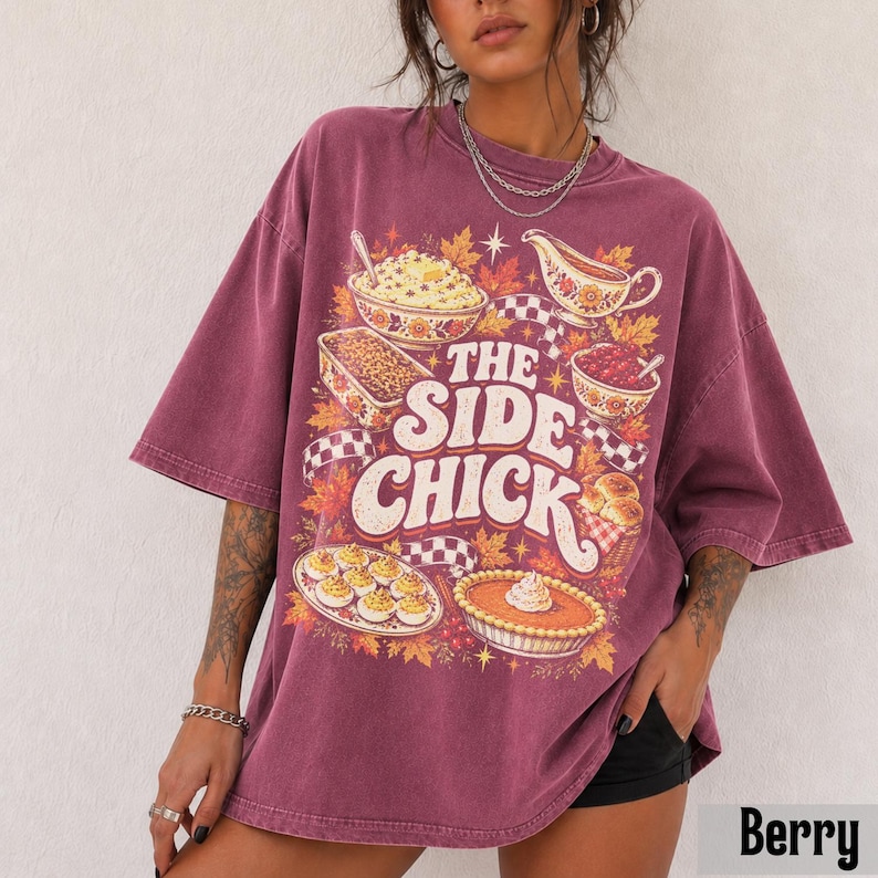 May include: A berry-colored oversized t-shirt featuring a graphic print of various food dishes, including a bowl of mashed potatoes, a casserole, a gravy boat, and a pie. The text reads THE SIDE CHICK in large, bold, cream-colored letters.