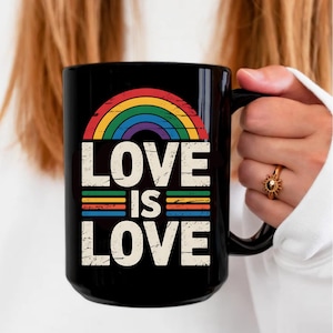 May include: Black ceramic mug with a rainbow design and the words "LOVE IS LOVE" in white block letters. The mug has a large handle and is held by a person. The rainbow design is at the top of the mug.