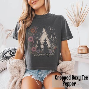May include: A dark gray cropped boxy tee featuring a graphic of two witch silhouettes in front of a bonfire, surrounded by flowers and the text "girls will be girls". The shirt is made of soft material.
