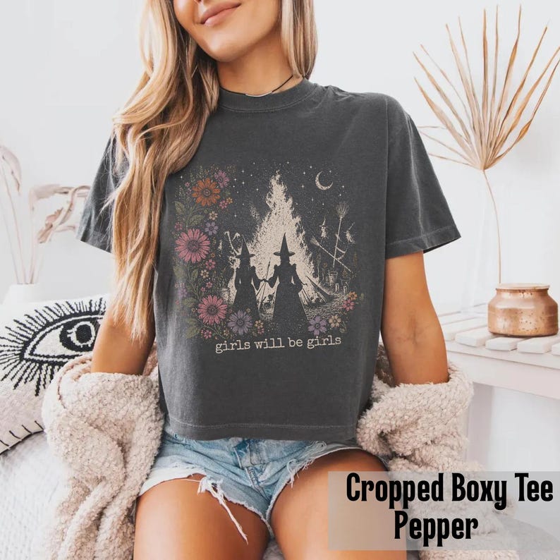May include: A dark gray cropped boxy tee featuring a graphic of two witch silhouettes in front of a bonfire, surrounded by flowers and the text "girls will be girls". The shirt is made of soft material.
