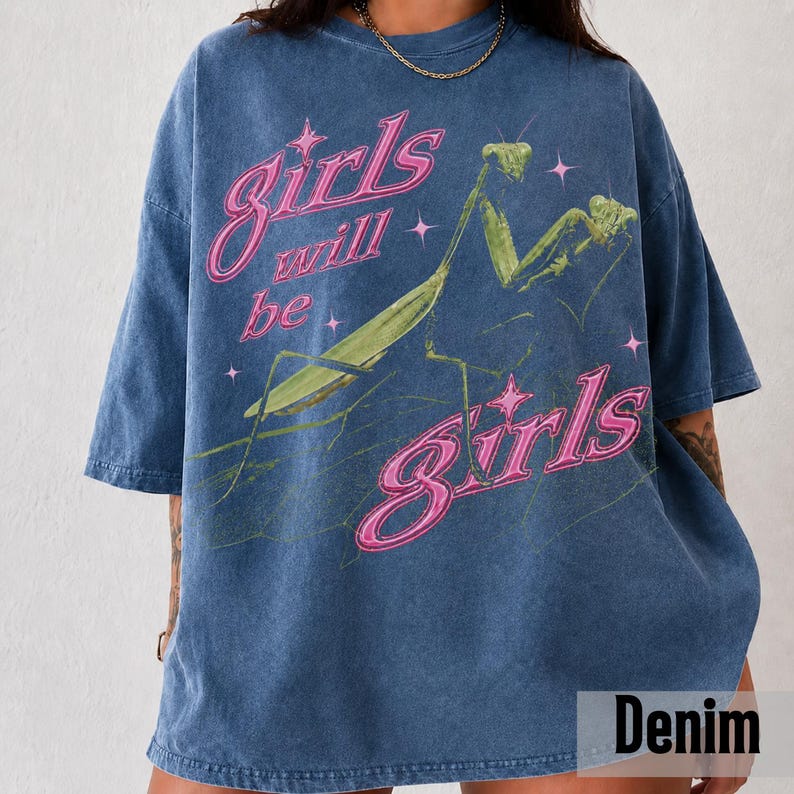 以下が含まれることがあります： 緑色のカマキリのグラフィックと、小さな星に囲まれたピンク色の「girls will be girls」という文字がプリントされた、ブルーのケミカルウォッシュ加工Tシャツ。半袖でクルーネックの、ゆったりとしたリラックスフィットのデザイン。