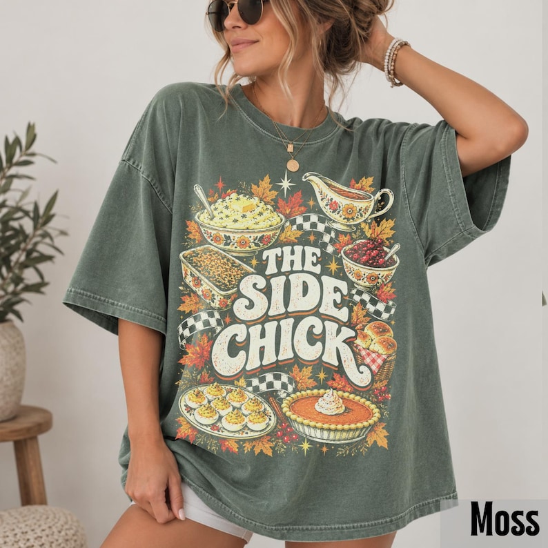 May include: A moss green oversized t-shirt featuring a graphic print of various Thanksgiving side dishes, including deviled eggs, a pie, and a casserole. The text reads THE SIDE CHICK in large white letters, surrounded by autumn leaves and checkered ribbons.