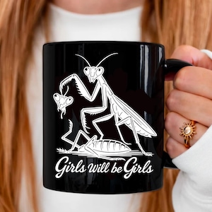 May include: Black ceramic mug featuring a white illustration of praying mantises. The design includes a large mantis, a smaller mantis, and a third insect, with the text "Girls will be Girls" in a cursive font.