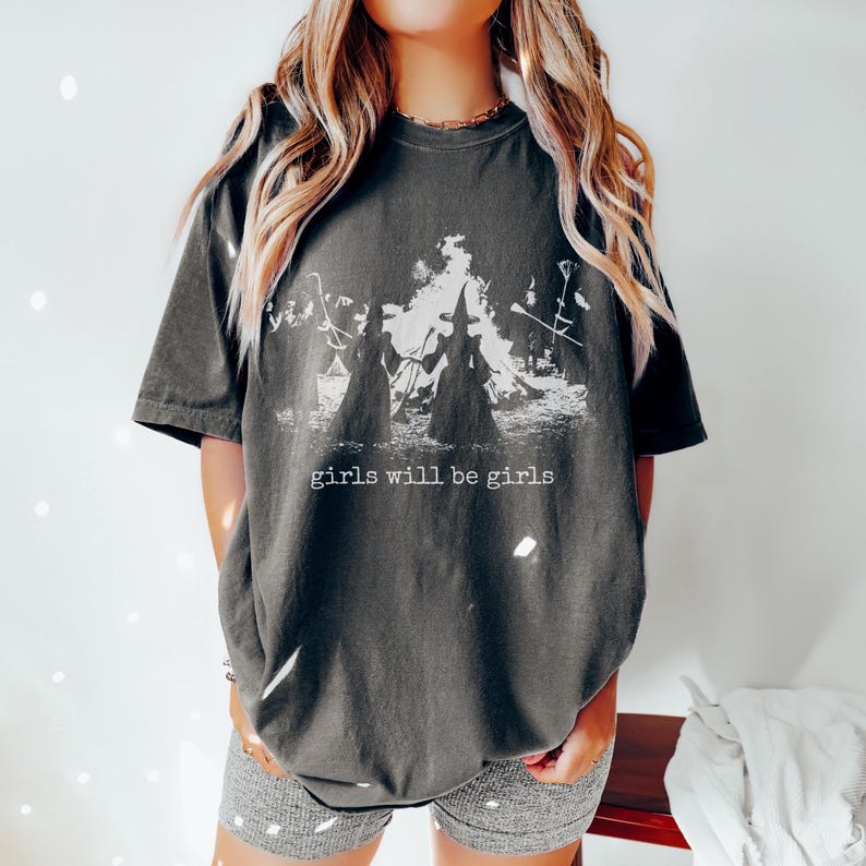 Pu&ograve; includere: Una t-shirt a maniche corte grigio antracite con una stampa grafica bianca di due streghe davanti a un fal&ograve;. Il testo "girls will be girls" &egrave; stampato in un carattere bianco stile macchina da scrivere sotto l'illustrazione.