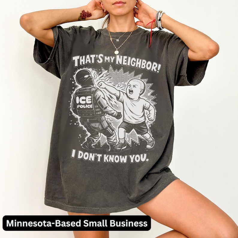 K&ouml;nnte beinhalten: Dunkelgraues T-Shirt mit einer Grafik, die ein Kind zeigt, das einen ICE-Polizisten schl&auml;gt. Der Text auf dem Shirt lautet: "THAT'S MY NEIGHBOR! I DON'T KNOW YOU." Das Shirt stammt von einem kleinen Unternehmen aus Minnesota.