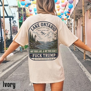 May include: An ivory t-shirt featuring a back graphic of Lake Ontario with a lighthouse, sailboat, and forest. Text includes "LAKE ONTARIO," "FRESHWATER. NOT FOR SALE," "PROTECT THE GREAT LAKES," "NOT YOUR LAKE. NOT YOUR LEGACY," and "FUCK TRUMP."