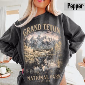 May include: A dark gray sweatshirt featuring a scenic illustration of the Grand Teton National Park. The design includes mountains, a lake, and trees, with the text "GRAND TETON NATIONAL PARK - EST 1929" in a vintage style. The sweatshirt has a crew neck.