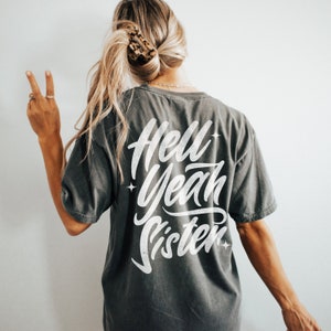 May include: A dark gray t-shirt with the text "Hell Yeah Sister" printed in white on the back. The text is in a stylized font with stars around it.