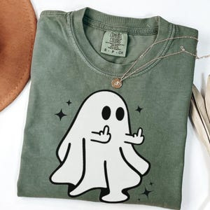 May include: Sage green t-shirt with a white ghost graphic giving the middle finger. The shirt has the words "Comfort Colors" on the tag. A gold necklace with a pendant is draped across the shirt.