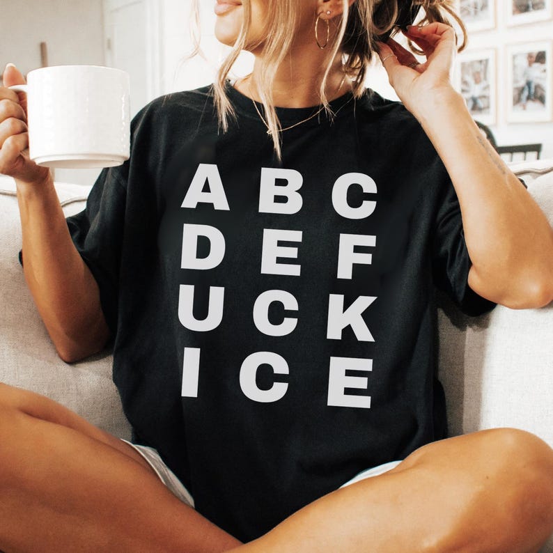 May include: Black t-shirt with the word "DUCK" spelled out vertically in white capital letters, with other letters arranged around it. The shirt is worn by a person holding a white mug.
