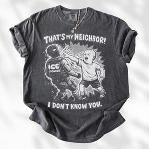 K&ouml;nnte beinhalten: Dunkelgraues T-Shirt mit einer grafischen Illustration eines Kindes, das einen ICE-Polizisten schl&auml;gt. Der Text auf dem Shirt lautet "THAT'S MY NEIGHBOR! I DON'T KNOW YOU."
