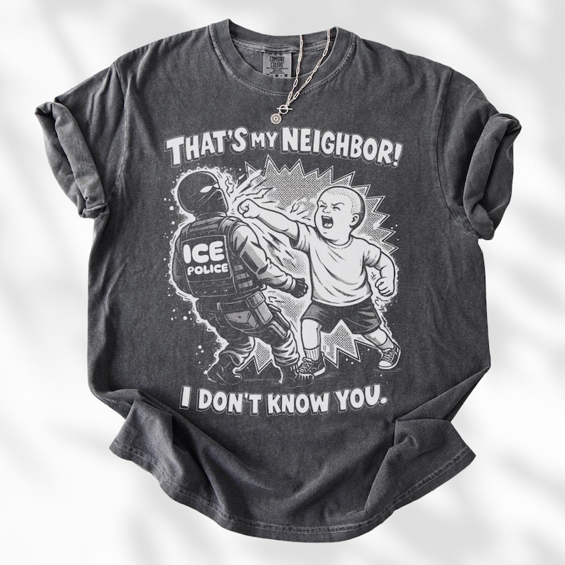 K&ouml;nnte beinhalten: Dunkelgraues T-Shirt mit einer grafischen Illustration eines Kindes, das einen ICE-Polizisten schl&auml;gt. Der Text auf dem Shirt lautet "THAT'S MY NEIGHBOR! I DON'T KNOW YOU."