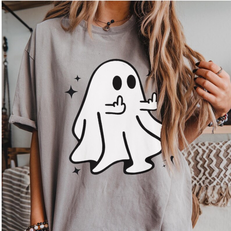 May include: Gray t-shirt featuring a white cartoon ghost giving the middle finger. The ghost has black eyes and is surrounded by four black star shapes. The shirt is a casual style.