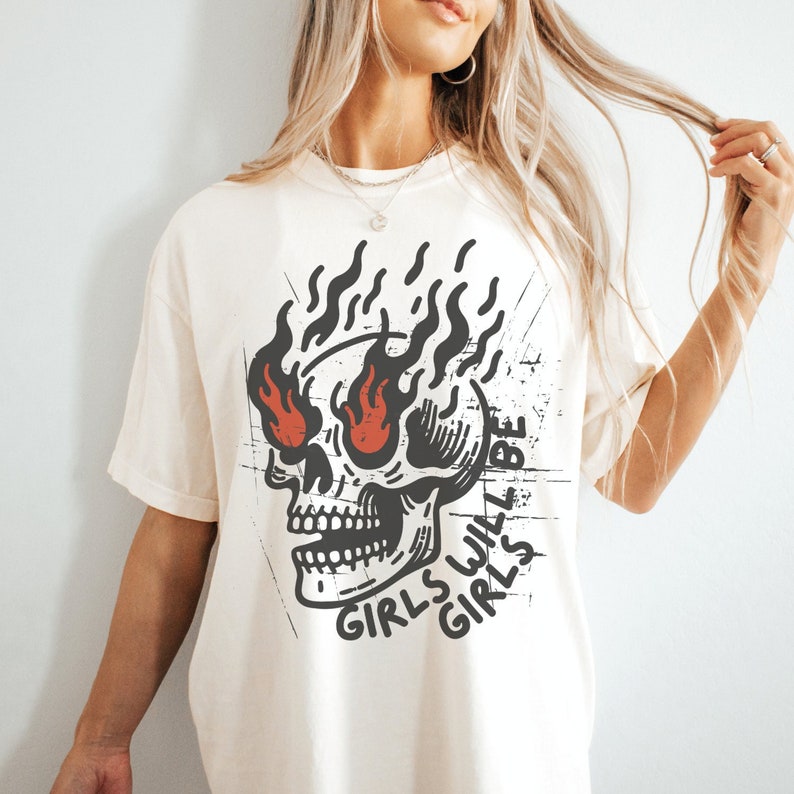 May include: White t-shirt featuring a black and red skull graphic with flames coming from its eyes. The text "Girls Will Be Girls" is written around the skull.