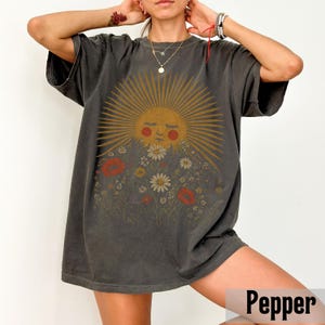Puede incluir: Camiseta gris oscuro con un dise&ntilde;o de sol sonriente rodeado de rayos y flores, incluyendo amapolas rojas y margaritas blancas. La camiseta tiene mangas cortas y un corte holgado.