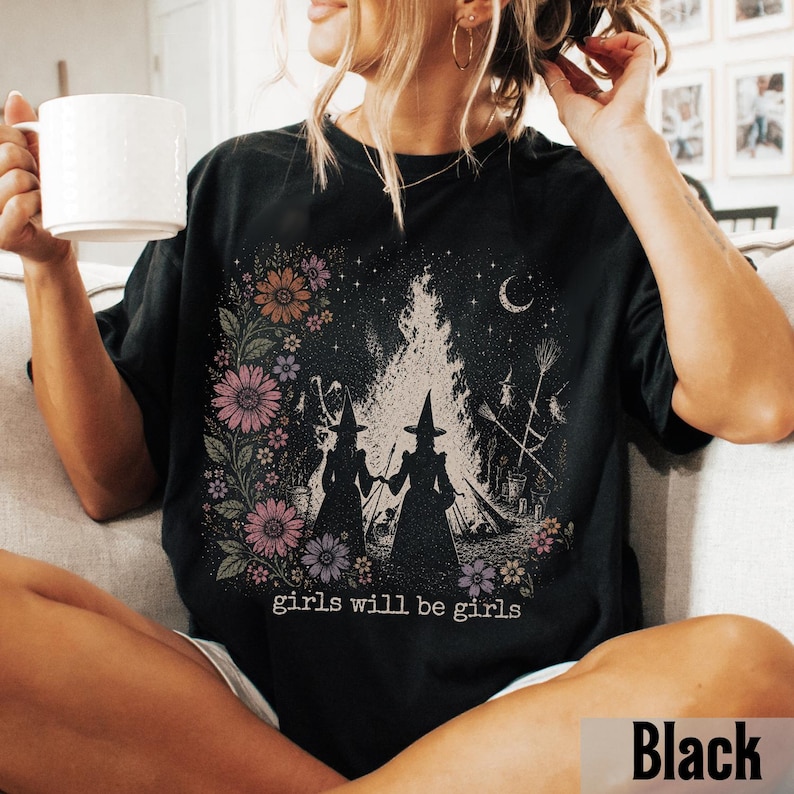 May include: Black t-shirt featuring a graphic of two witches holding hands in front of a bonfire, surrounded by flowers and a crescent moon. The text "girls will be girls" is printed below the graphic. The shirt is a relaxed fit.
