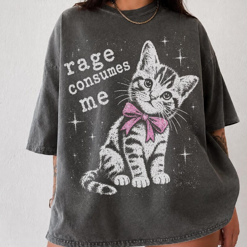 May include: A charcoal gray short-sleeve t-shirt featuring a white graphic of a tabby kitten wearing a pink bow. The text "rage consumes me" is printed in white next to the kitten, surrounded by small white star shapes across the front.