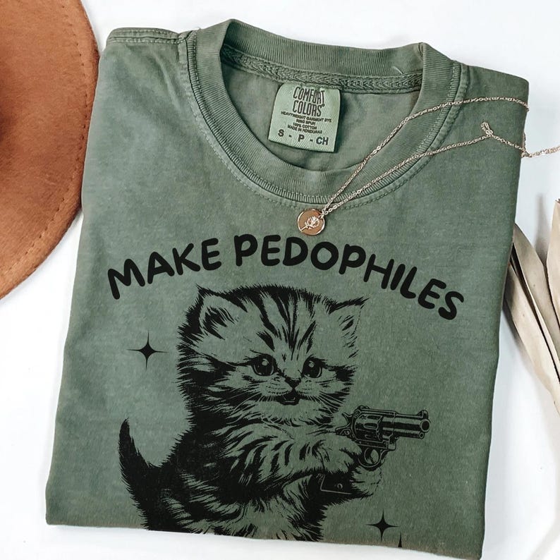 Op de afbeelding: Olijfgroen t-shirt met de tekst "MAKE PEDOPHILES" boven een cartoonkatje met een pistool. Het shirt heeft een ronde hals en korte mouwen. Een zilveren ketting met een hanger is over het shirt gedrapeerd.