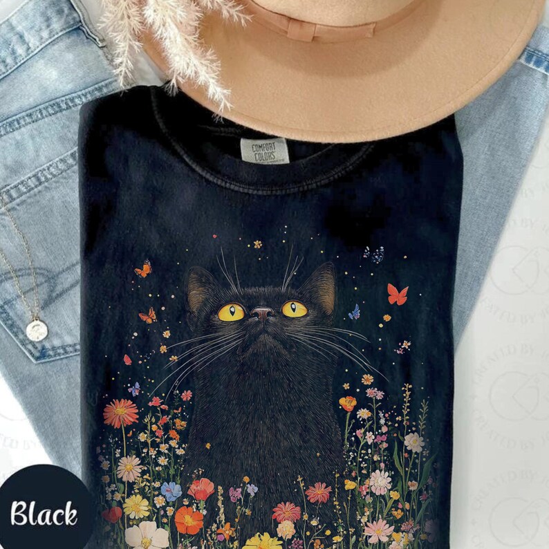 May include: Black t-shirt with a black cat with yellow eyes surrounded by colorful flowers and butterflies. The shirt has the text "Comfort Colors" on the tag.