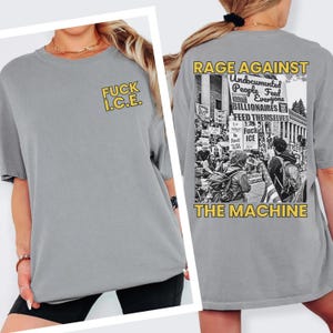 Puede incluir: Una camiseta gris con el texto "FUCK I.C.E." en amarillo en la parte delantera. La parte trasera de la camiseta presenta una imagen en blanco y negro de una protesta con el texto "RAGE AGAINST THE MACHINE".