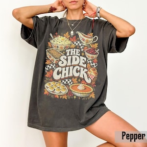 May include: A charcoal gray short-sleeve t-shirt featuring a graphic print of various Thanksgiving dishes, including a pie, deviled eggs, and a gravy boat, with the text THE SIDE CHICK in large white letters surrounded by autumn leaves.