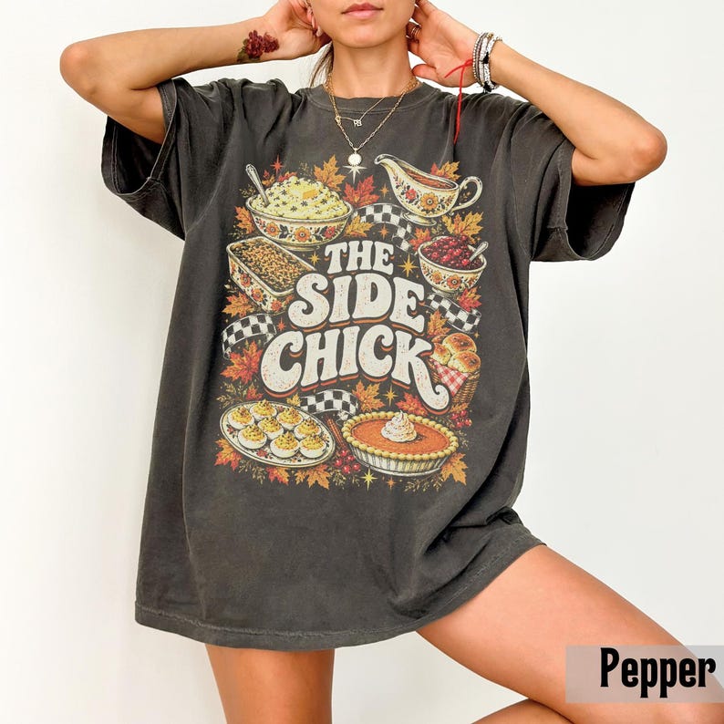 May include: A charcoal gray short-sleeve t-shirt featuring a graphic print of various Thanksgiving dishes, including a pie, deviled eggs, and a gravy boat, with the text THE SIDE CHICK in large white letters surrounded by autumn leaves.