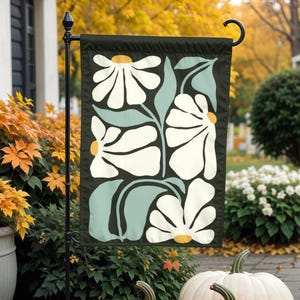 Puede incluir: Una bandera de jardín con un diseño floral estilizado. La bandera presenta flores blancas con centros amarillos sobre un fondo verde oscuro.
