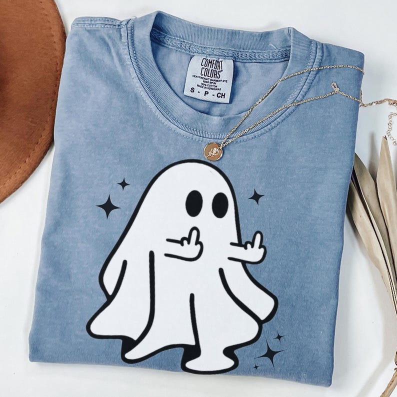 May include: A blue Comfort Colors t-shirt featuring a white cartoon ghost giving the middle finger, with black star accents. A gold necklace with a pendant is draped across the shirt.