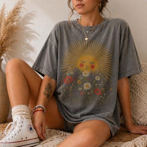 Puede incluir: Una camiseta gris con un dise&ntilde;o de sol sonriente rodeado de flores coloridas. El sol tiene rayos. La camiseta tiene mangas cortas y un corte holgado, ideal para un look casual.