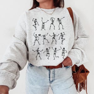 K&ouml;nnte beinhalten: Hellgraues Sweatshirt mit einer schwarzen Illustration tanzender Skelette. Die Skelette sind in verschiedenen Posen dargestellt. Das Sweatshirt hat lange &Auml;rmel und einen Rundhalsausschnitt.