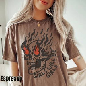 May include: A brown t-shirt featuring a graphic of a skull with flames and the text "Girls will be girls".