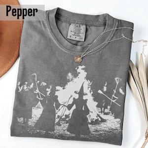 Pu&ograve; includere: Una t-shirt color pepe con una stampa grafica bianca di figure con cappelli a punta vicino a un fal&ograve;. La maglietta ha un girocollo e un'etichetta Comfort Colors all'interno. Una sottile collana a catena color oro &egrave; appoggiata sopra.