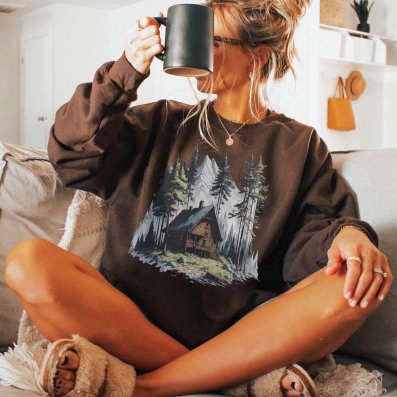 May include: A brown sweatshirt featuring a mountain cabin scene with trees and a mountain range. The sweatshirt is a dark brown color. The person is holding a black mug.