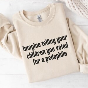 May include: A beige crewneck sweatshirt with the text "imagine telling your children you voted for a pedophile" in black. The sweatshirt is folded and displayed on a white surface.
