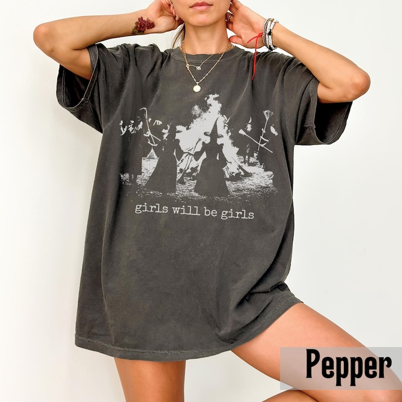 May include: Dark grey t-shirt with a graphic print of witches around a bonfire, with the text "girls will be girls" in a white font. The shirt has short sleeves and a relaxed fit.