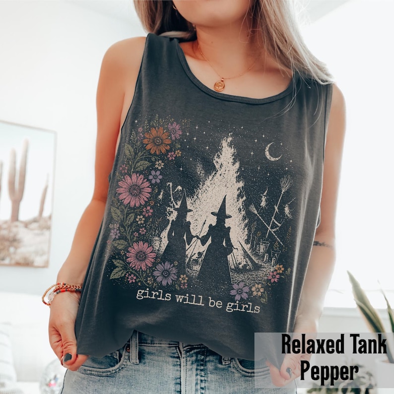 May include: A dark gray relaxed tank top with a floral and witch-themed graphic. The design features two witch silhouettes, a bonfire, flowers, and the text "girls will be girls". The tank top is a pepper color.