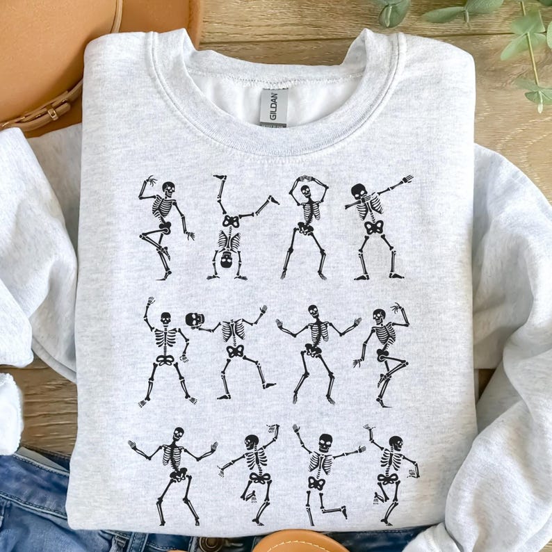 K&ouml;nnte beinhalten: Ein hellgraues Sweatshirt mit einem sich wiederholenden Muster aus schwarzen Skelettfiguren in verschiedenen Tanzposen. Die Skelette sind detailliert mit sichtbaren Brustk&ouml;rben, Sch&auml;deln und Gliedma&szlig;en, was ein verspieltes und festliches Design ergibt.