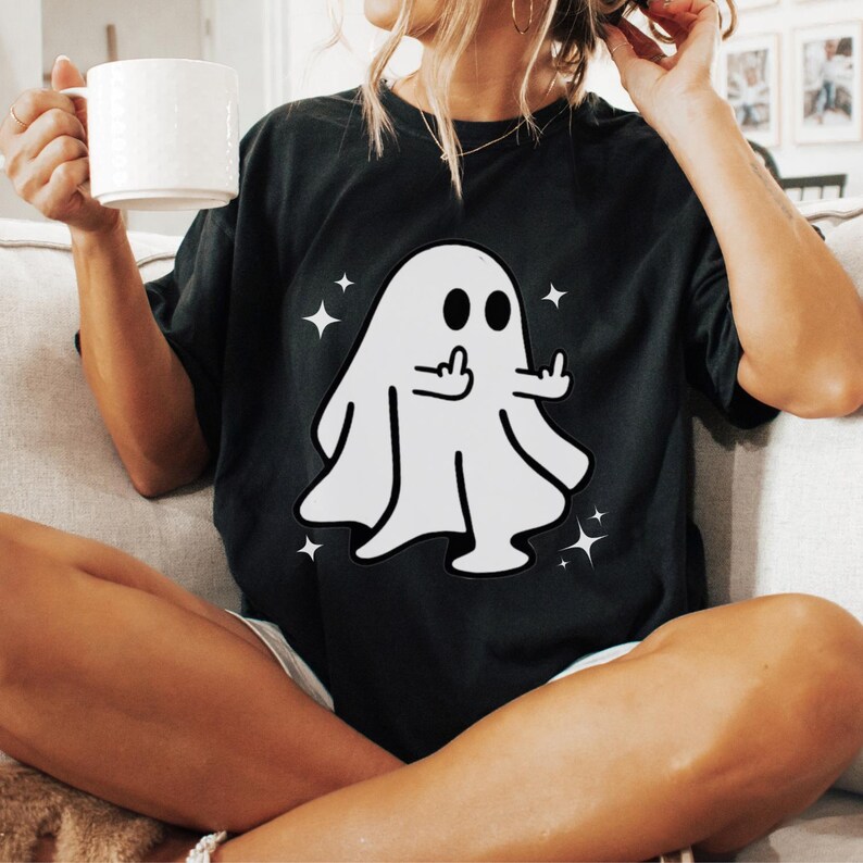 May include: Black t-shirt featuring a white cartoon ghost giving the middle finger, surrounded by star graphics. The ghost has two black eyes and a simple outline. A casual, humorous design.