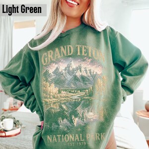 May include: Light green sweatshirt with a graphic of the Grand Teton National Park. The design features a mountain range, lake, and trees, with the words "GRAND TETON NATIONAL PARK EST 1929" in gold lettering.