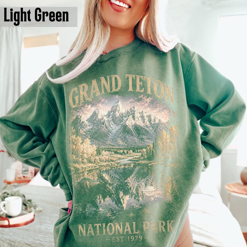 May include: Light green sweatshirt with a graphic of the Grand Teton National Park. The design features a mountain range, lake, and trees, with the words "GRAND TETON NATIONAL PARK EST 1929" in gold lettering.