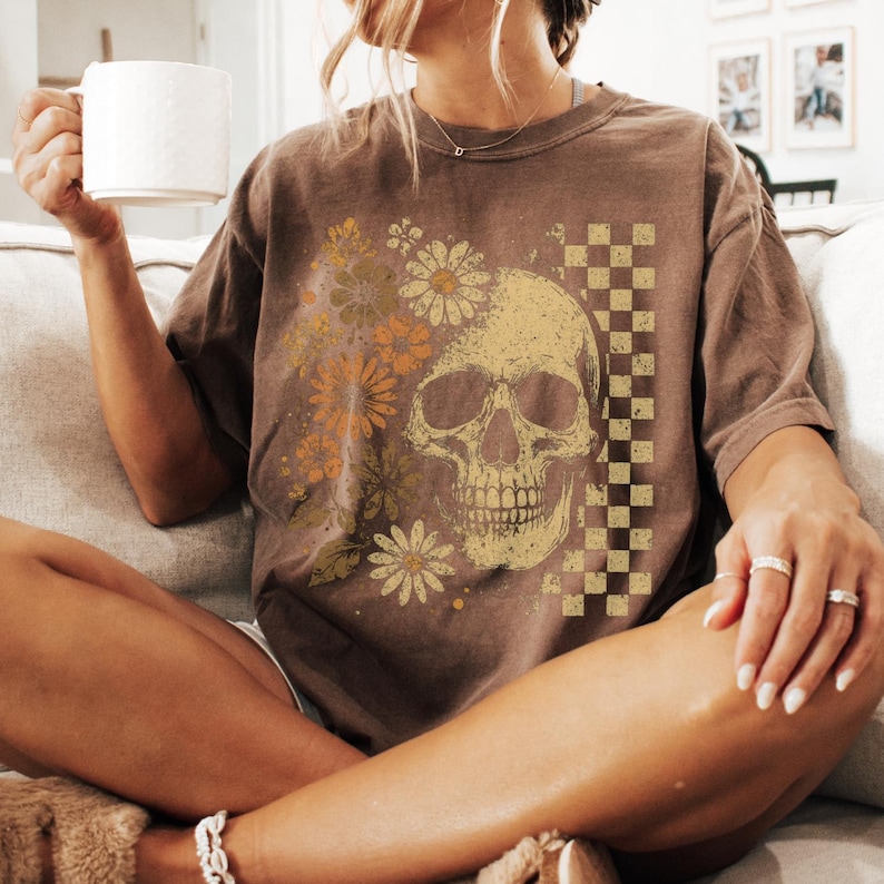 May include: Brown t-shirt featuring a skull design with floral and checkered accents. The skull is in a vintage style, with flowers in shades of yellow and orange. The shirt has a relaxed fit.