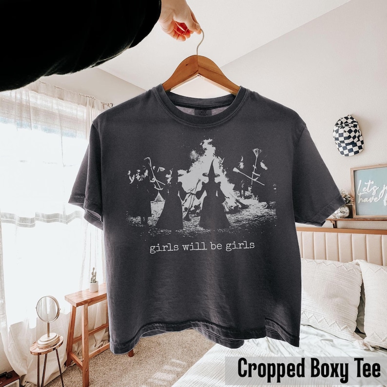 Pu&ograve; includere: Una t-shirt a maniche corte grigio antracite con una stampa grafica bianca di tre figure con cappelli a punta davanti a un grande fal&ograve;. Il testo "girls will be girls" &egrave; stampato in bianco sotto la grafica sul davanti della maglietta.