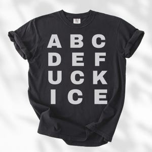 May include: Black t-shirt with the word "DUCK" spelled out in large, white, block letters. The letters "A, B, C, D, E, F, U, C, K, I, C, E" are arranged in a grid pattern. The shirt has rolled-up sleeves.