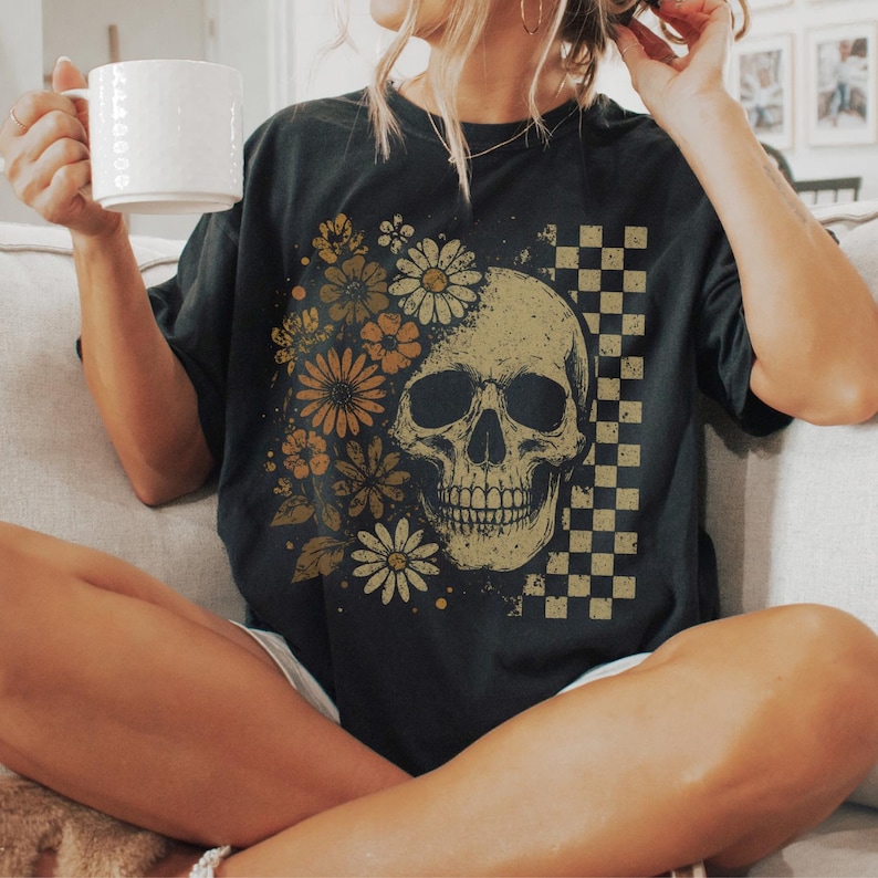 May include: Black t-shirt featuring a skull design with floral accents and a checkered pattern. The skull is in the center, with flowers on the left side. The shirt is a dark color and has a vintage look.