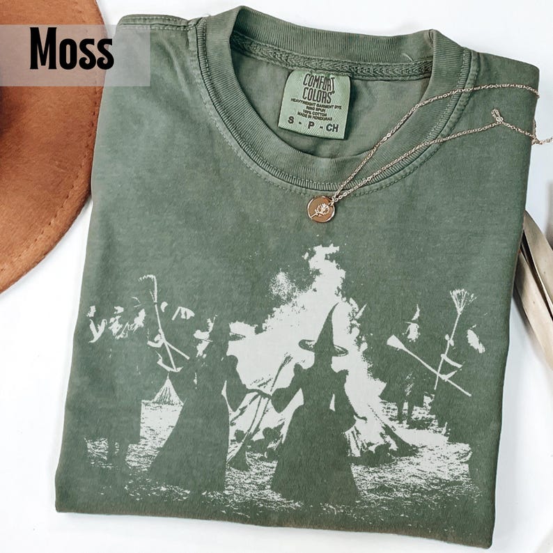 Pu&ograve; includere: Una t-shirt a maniche corte color verde muschio con una stampa grafica bianca di figure con cappelli a punta che tengono scope vicino a un fal&ograve;. La maglietta ha un girocollo e un'etichetta del marchio Comfort Colors all'interno del colletto.