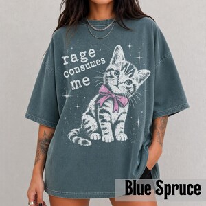 May include: A dark blue-grey oversized t-shirt featuring a white graphic of a kitten wearing a pink bow. The text "rage consumes me" is printed in white above the kitten, surrounded by small white starburst accents across the front of the shirt.
