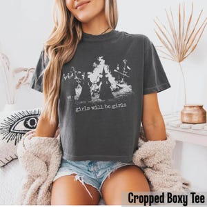Pu&ograve; includere: Una t-shirt a maniche corte color grigio antracite con una stampa grafica bianca raffigurante sagome di streghe attorno a un fal&ograve;. Il testo "girls will be girls" &egrave; stampato in bianco sotto la grafica. Il capo ha un taglio corto e squadrato.